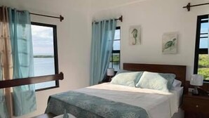 A bedroom with a bed, curtains, a bedside table, and a view of the outdoors. - Bay Towers Hotel (Caye Caulker)
