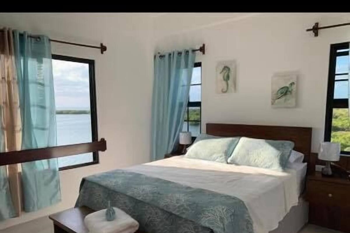 Een slaapkamer met een bed, gordijnen, een nachtkastje en uitzicht op het omliggende landschap.