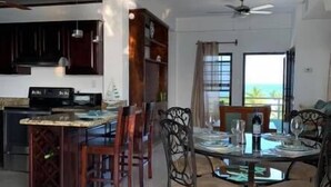 A modern kitchen with dark cabinetry, a dining area with a glass table and chairs, and a view of the ocean through large windows. - Bay Towers Hotel (Caye Caulker)