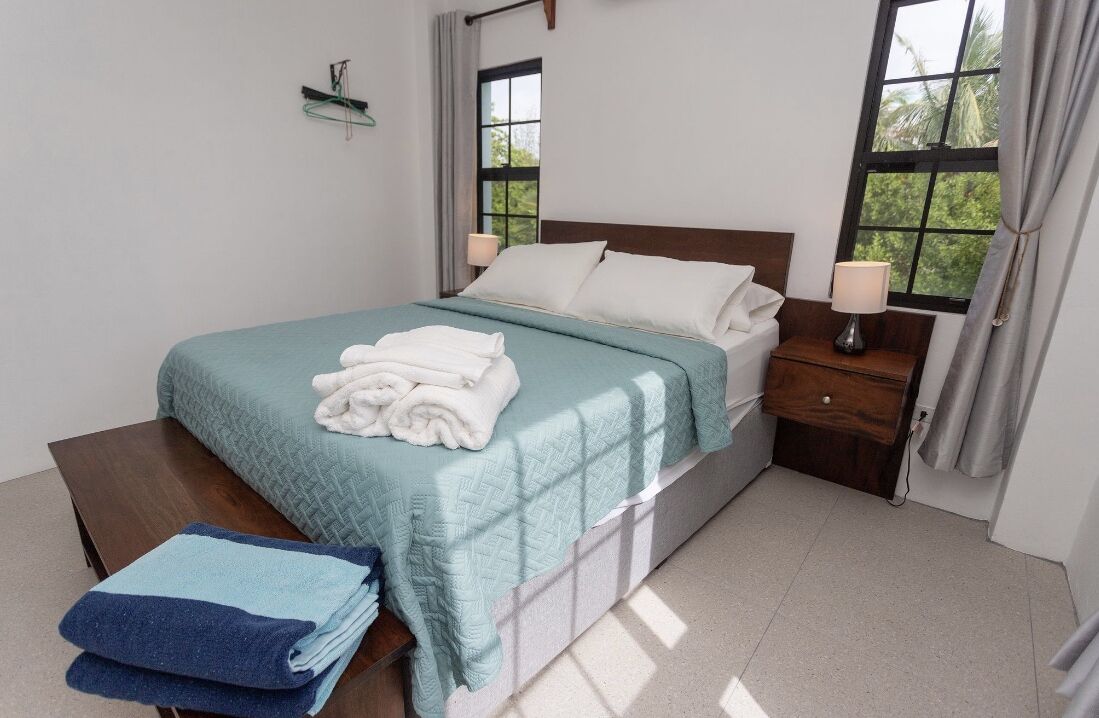 A neatly made bed with a teal bedspread and white pillows, a wooden nightstand with a lamp, and a window with curtains.