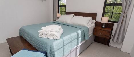 A neatly made bed with a teal bedspread and white pillows, a wooden nightstand with a lamp, and a window with curtains.