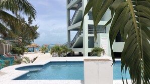 A pool area with a white pool, palm trees, and a building with balconies. - Bay Towers Hotel (Caye Caulker)