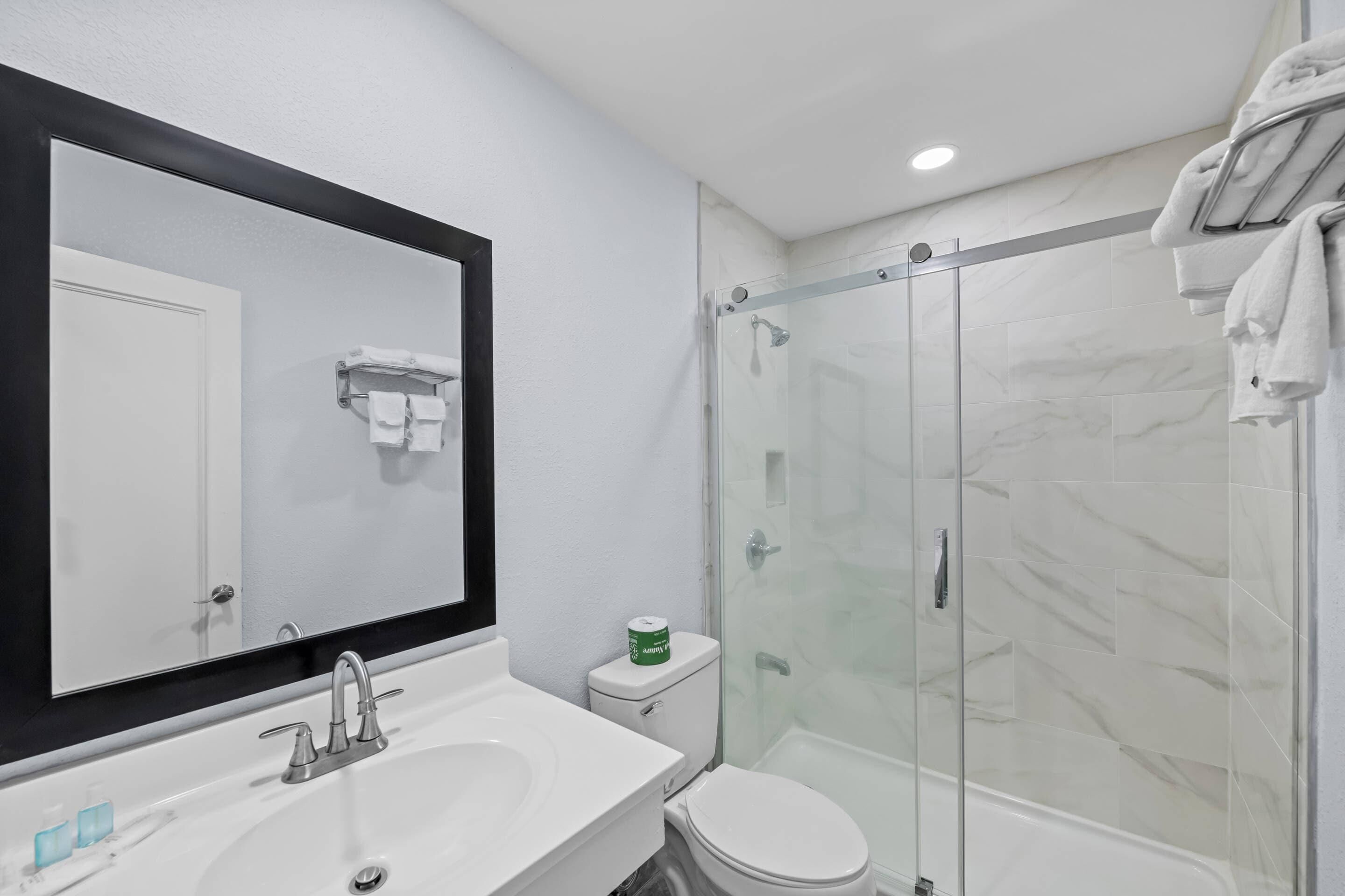 Un baño moderno con un espejo grande, lavamanos blanco, inodoro y ducha con mampara de vidrio.