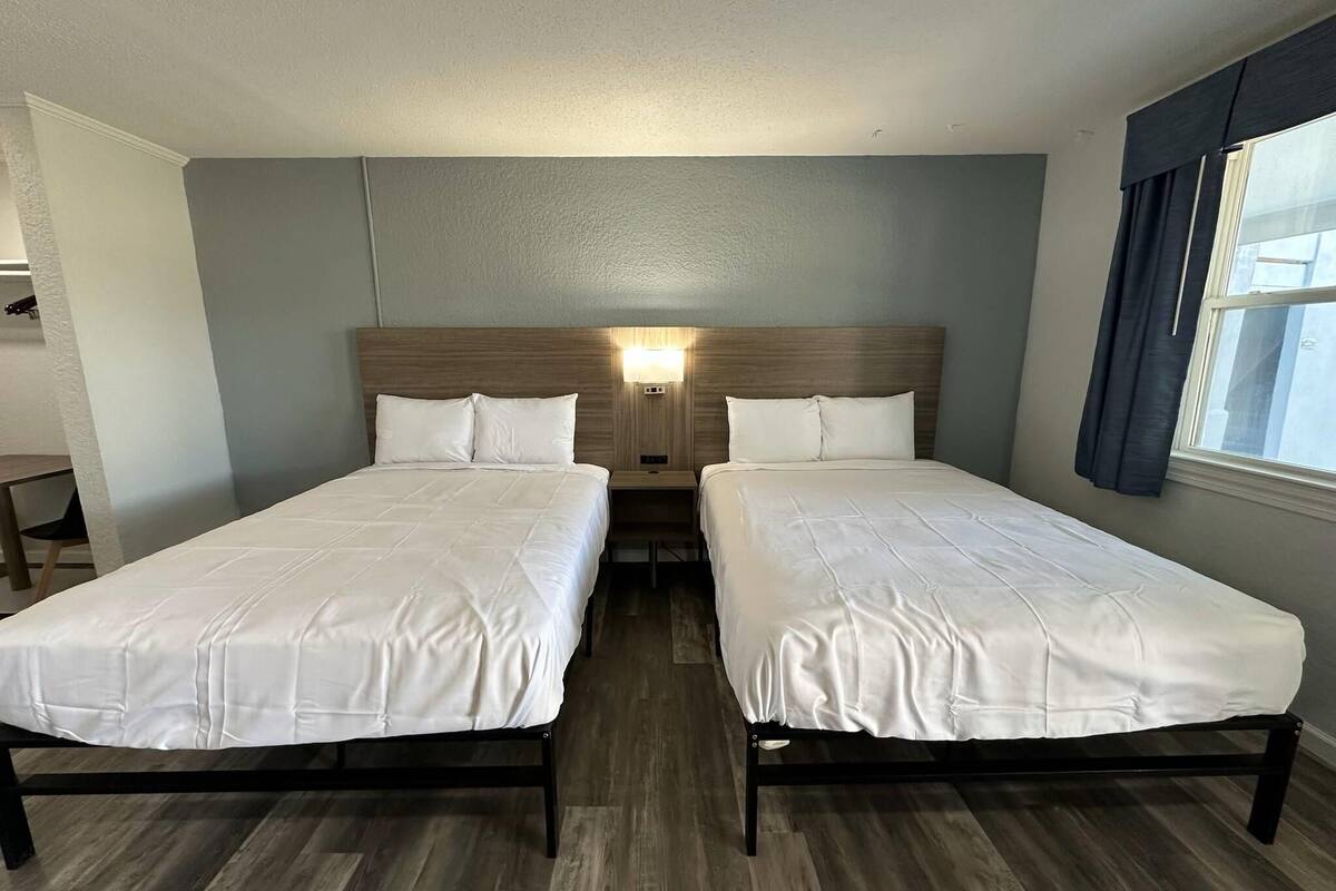 Een hotelkamer met twee bedden, een houten hoofdbord, een nachtkastje met een lamp en een raam met gordijnen.