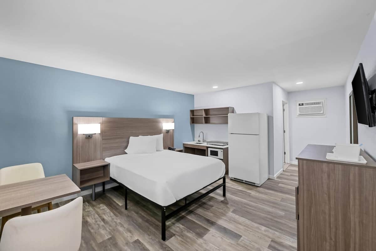 Een hotelkamer met een bed, nachtkastje, lamp, bureau, stoel en kitchenette.