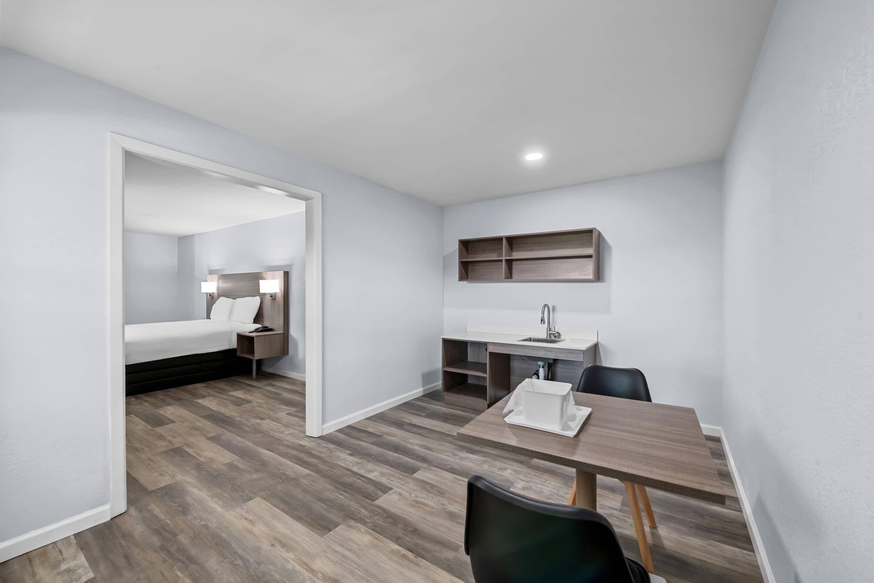 A compact room with a bed, a desk with a chair, a small sink, and a shelf.