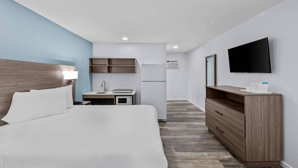 A hotel room with a bed, a desk with a lamp, a TV mounted on the wall, and a kitchenette area.