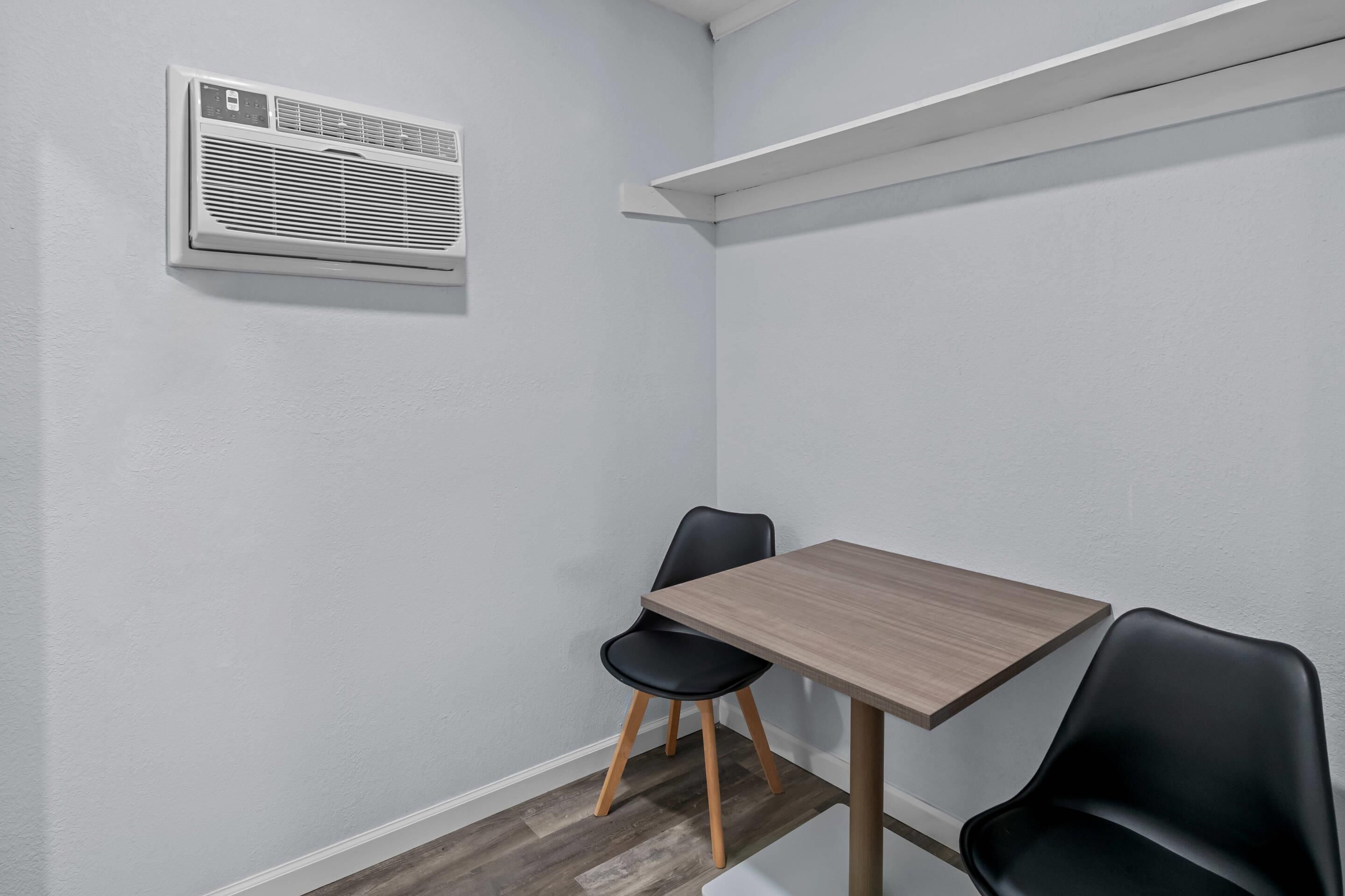Quarto de canto com uma mesa de madeira, duas cadeiras pretas, um ar condicionado branco e uma prateleira branca.