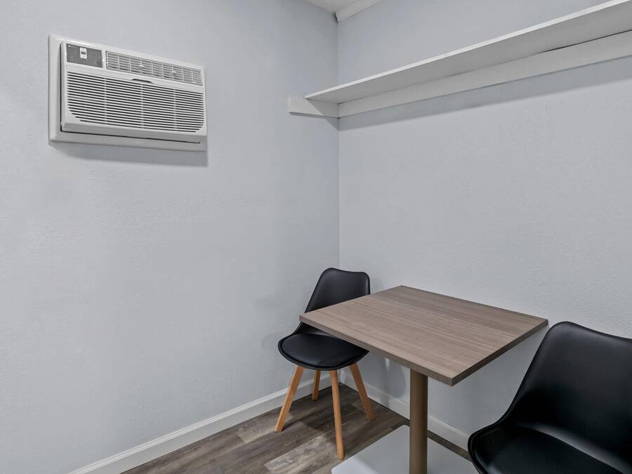 Quarto de canto com uma mesa de madeira, duas cadeiras pretas, um ar condicionado branco e uma prateleira branca.