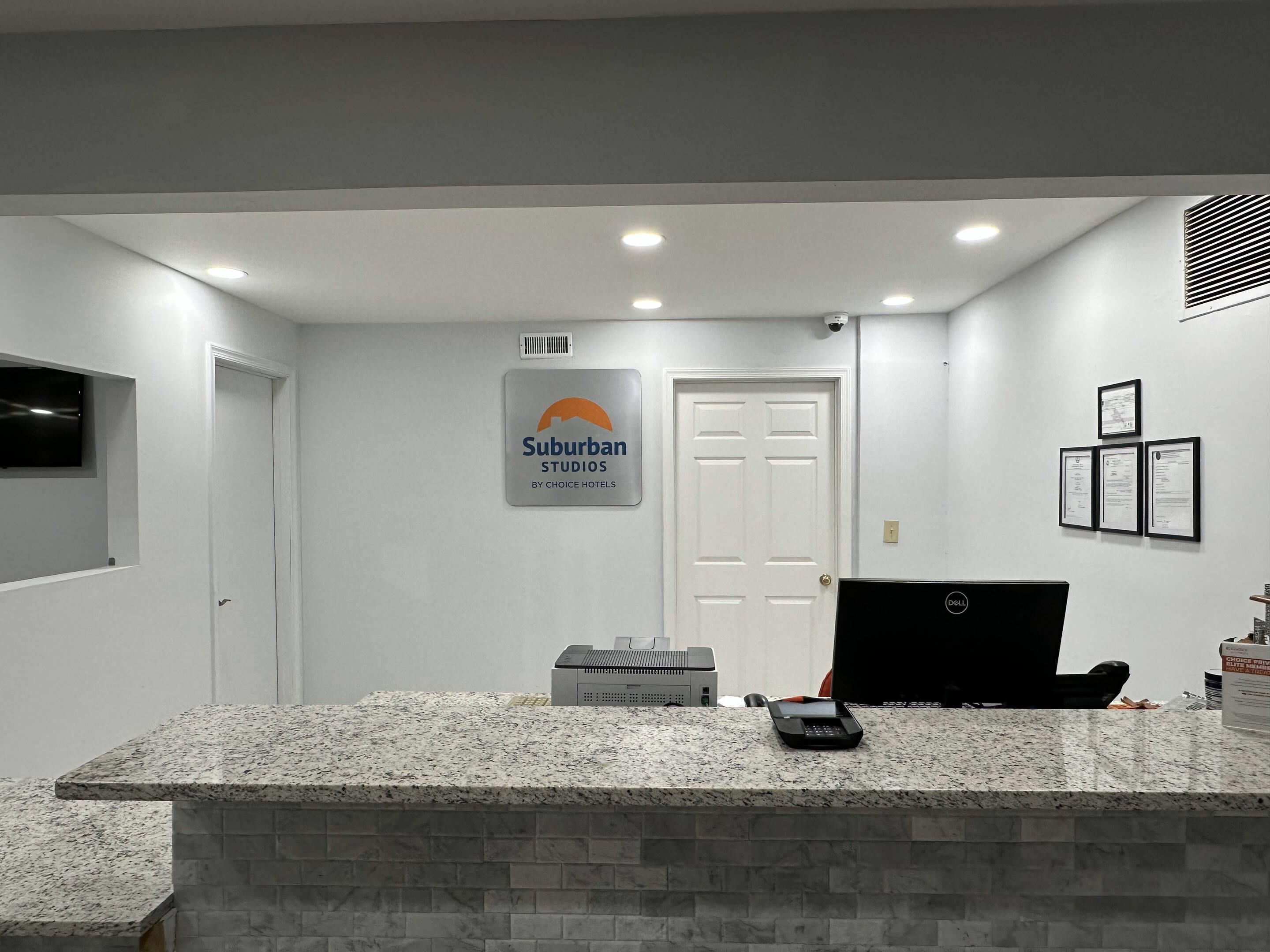 A reception area with a sign for Suburban Studios by Choice Hotels, a desktop computer, a telephone, and a small trash bin.