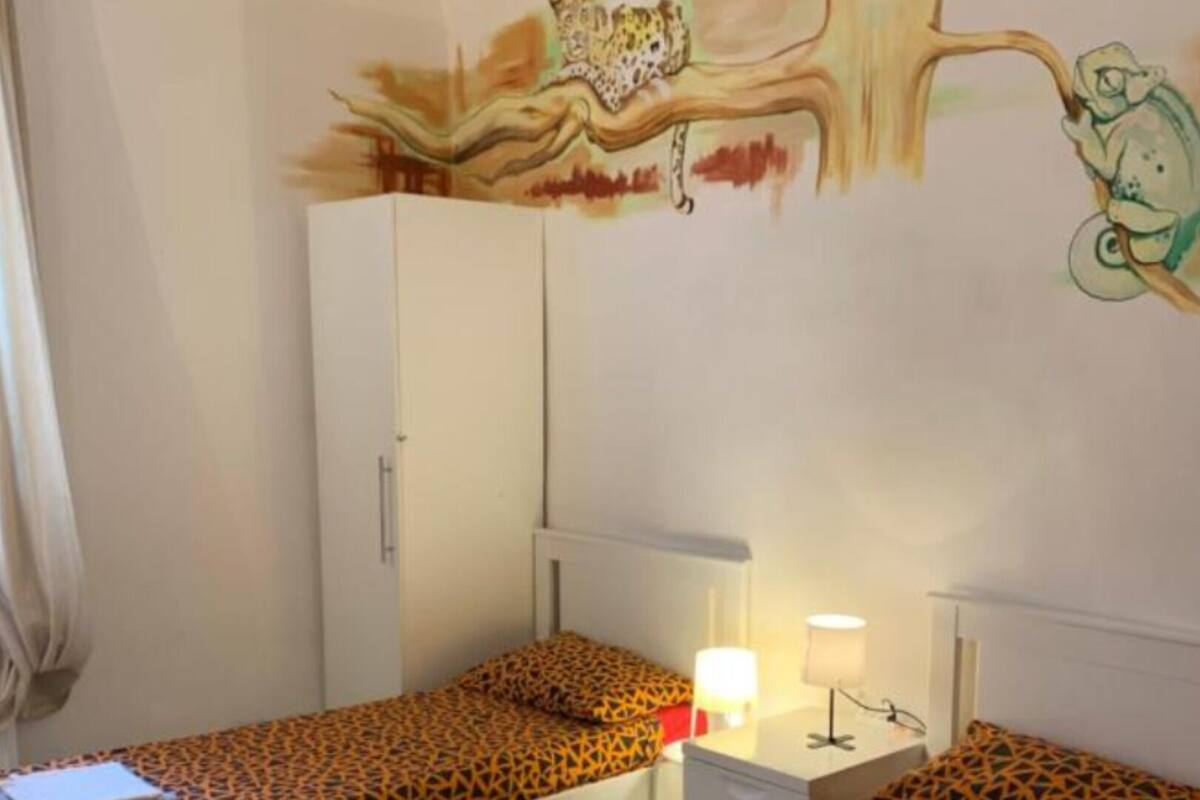 A room with two single beds, a wardrobe, a desk with a lamp, and a wall mural.
