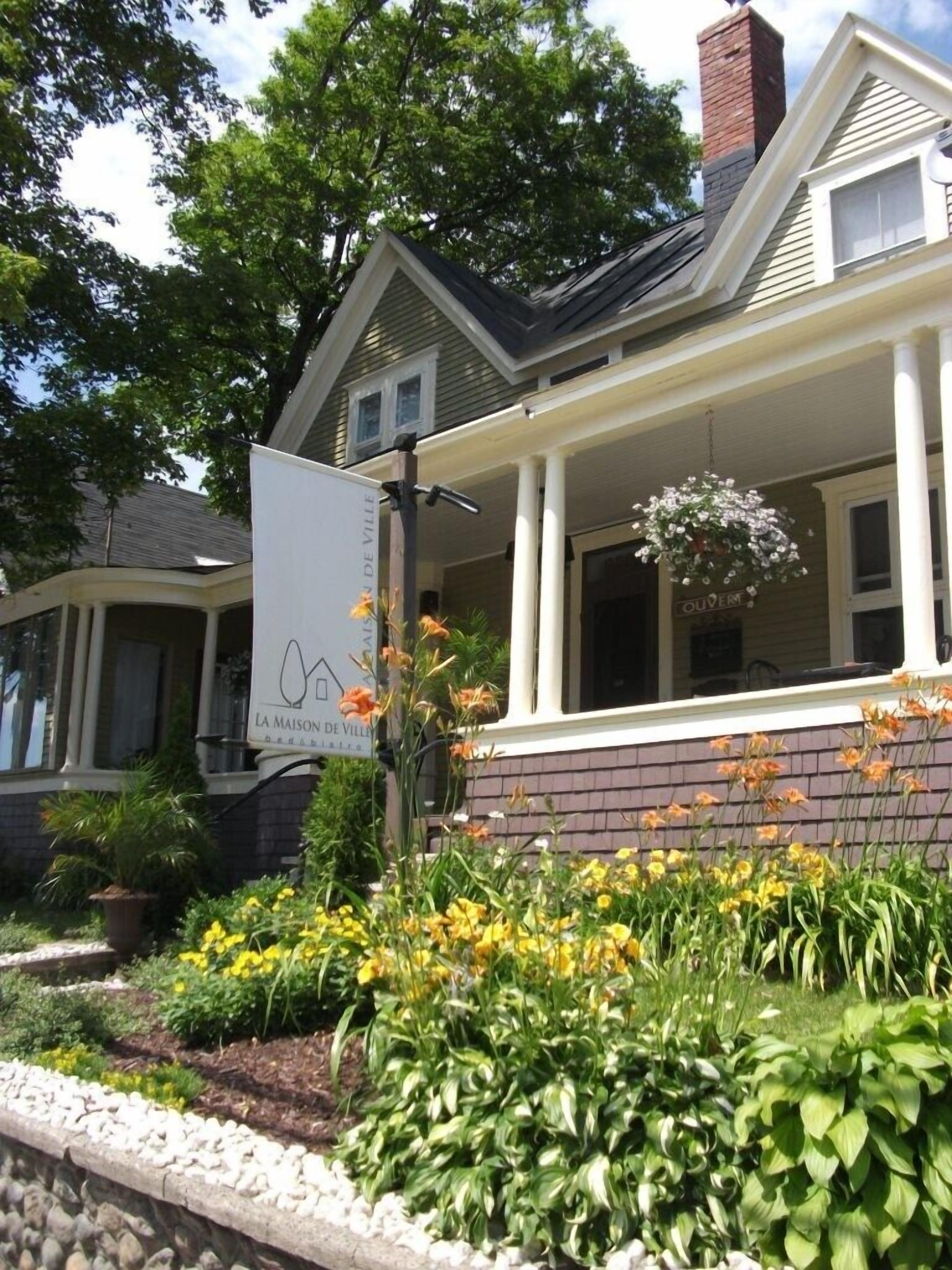 A two-story house with a porch, a sign reading "La Maison de Ville," and a variety of flowers in the front yard.