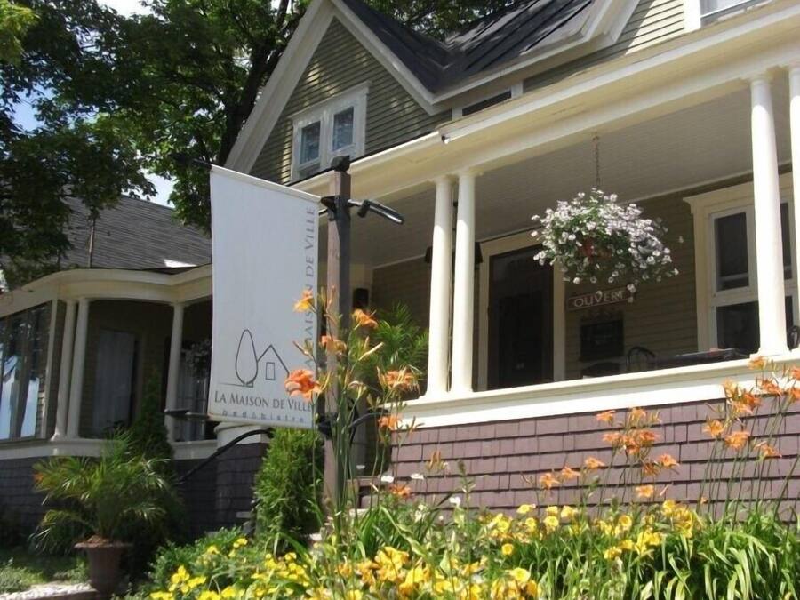 A two-story house with a porch, a sign reading "La Maison de Ville," and a variety of flowers in the front yard.