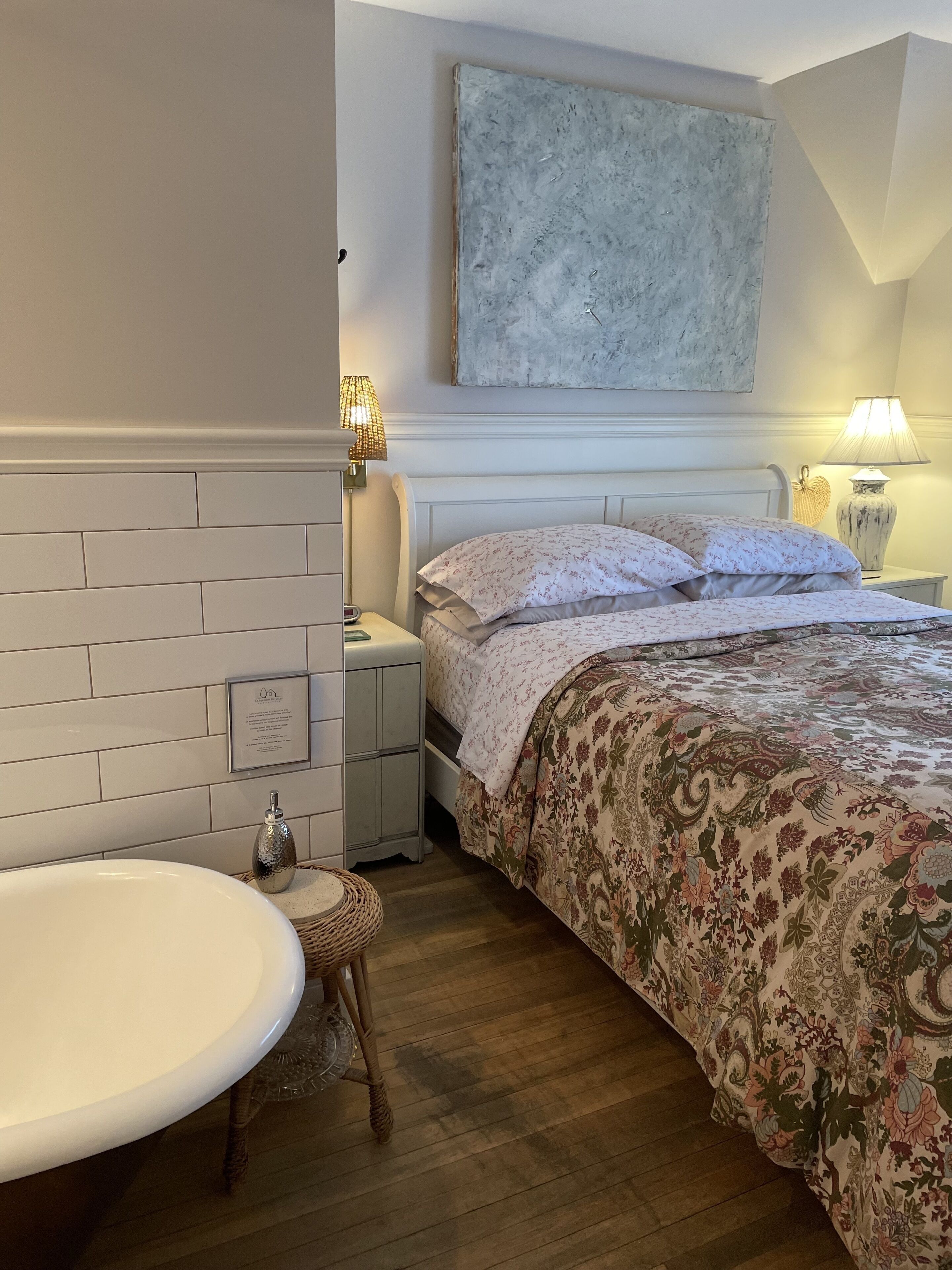 A bedroom with a white brick wall, a large bed with floral bedding, a nightstand with a lamp, and a white sink.