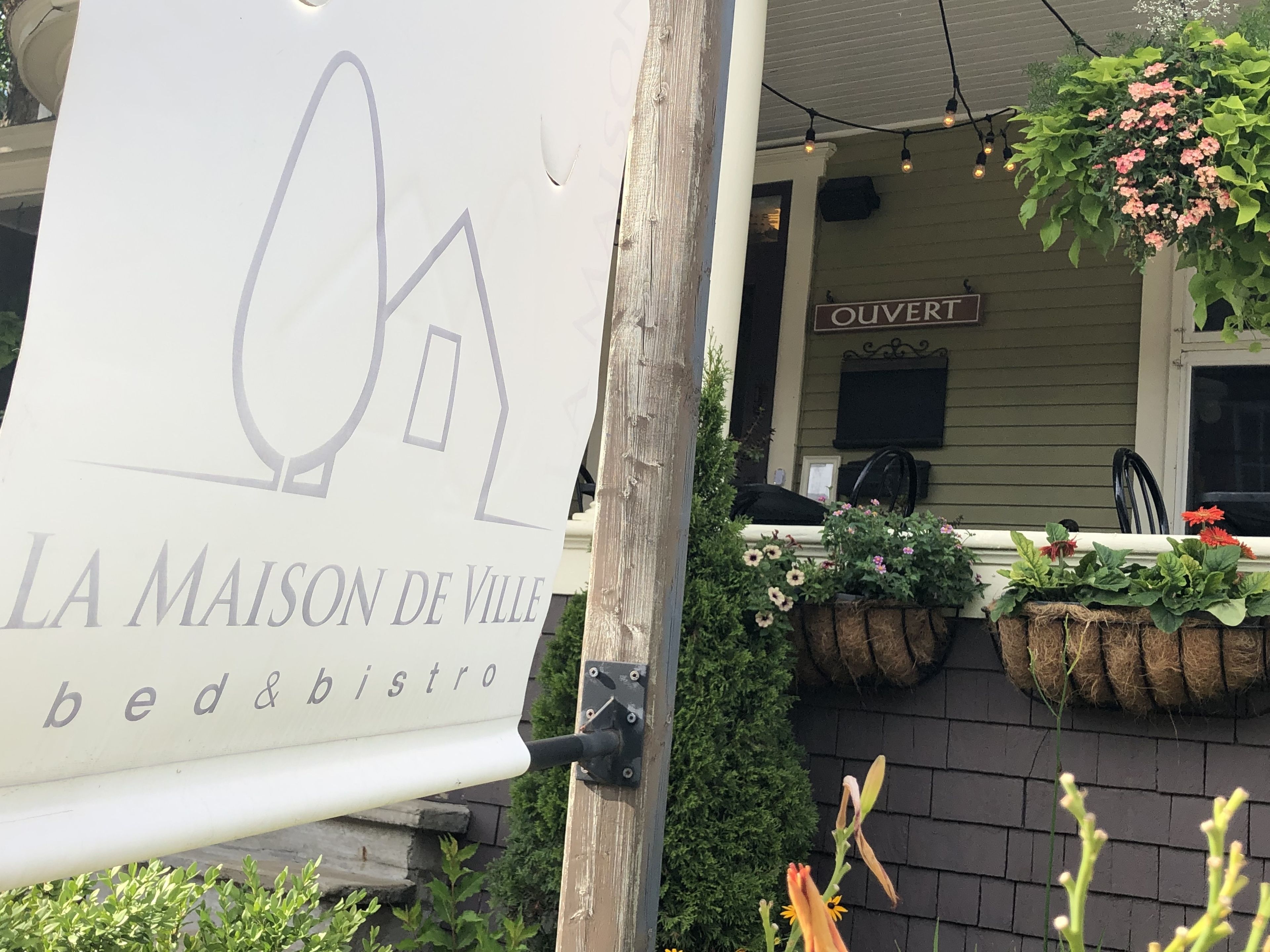 Letrero de 'La Maison de Ville bed & bistro' con el logo de una casa, un porche con cestas de flores colgantes y un letrero que dice 'OUVERT'.