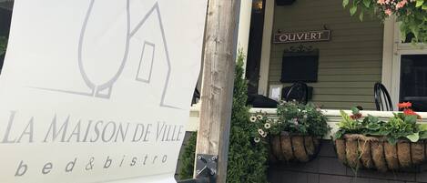 Letrero de 'La Maison de Ville bed & bistro' con el logo de una casa, un porche con cestas de flores colgantes y un letrero que dice 'OUVERT'.