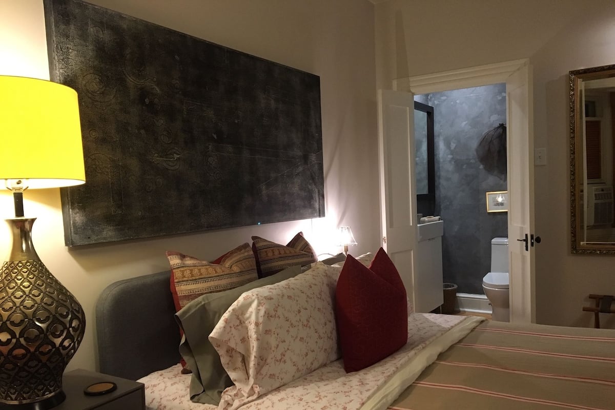 Een slaapkamer met een bed, een nachtkastje, een lamp, een groot abstract schilderij en een deur die naar een badkamer leidt.