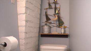 Un baño con un inodoro blanco, un rollo de papel higiénico, un bote de basura y una escultura metálica decorativa en un estante sobre el inodoro.