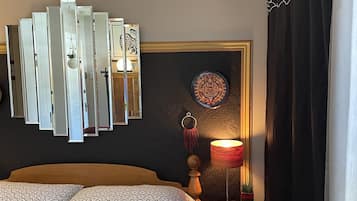 A bedroom with a bed, a nightstand with a lamp, a wall-mounted mirror, and a decorative wall piece.