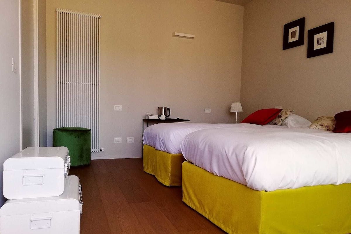 Une chambre d’hôtel avec deux lits, un radiateur, une poubelle verte, une petite table avec une bouilloire et deux valises blanches.