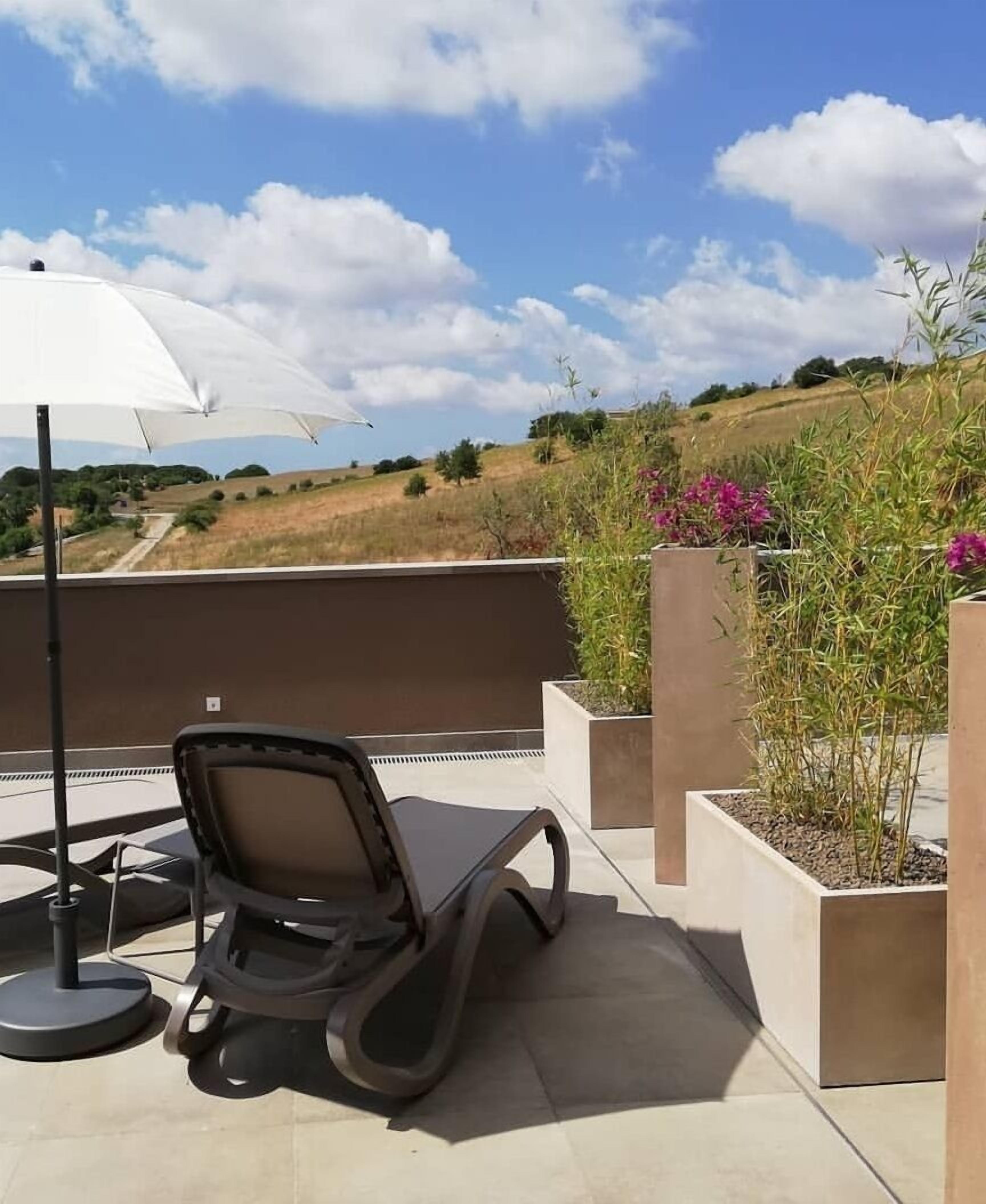 A rooftop terrace with a lounge chair, an umbrella, and planters with flowers.