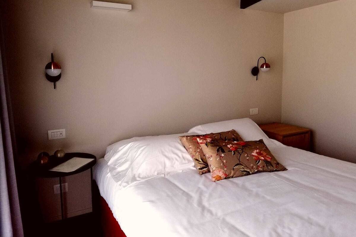 Un lit bien fait, avec du linge de lit blanc et un coussin à motifs floraux, une table de chevet avec une lampe et un luminaire fixé au mur.