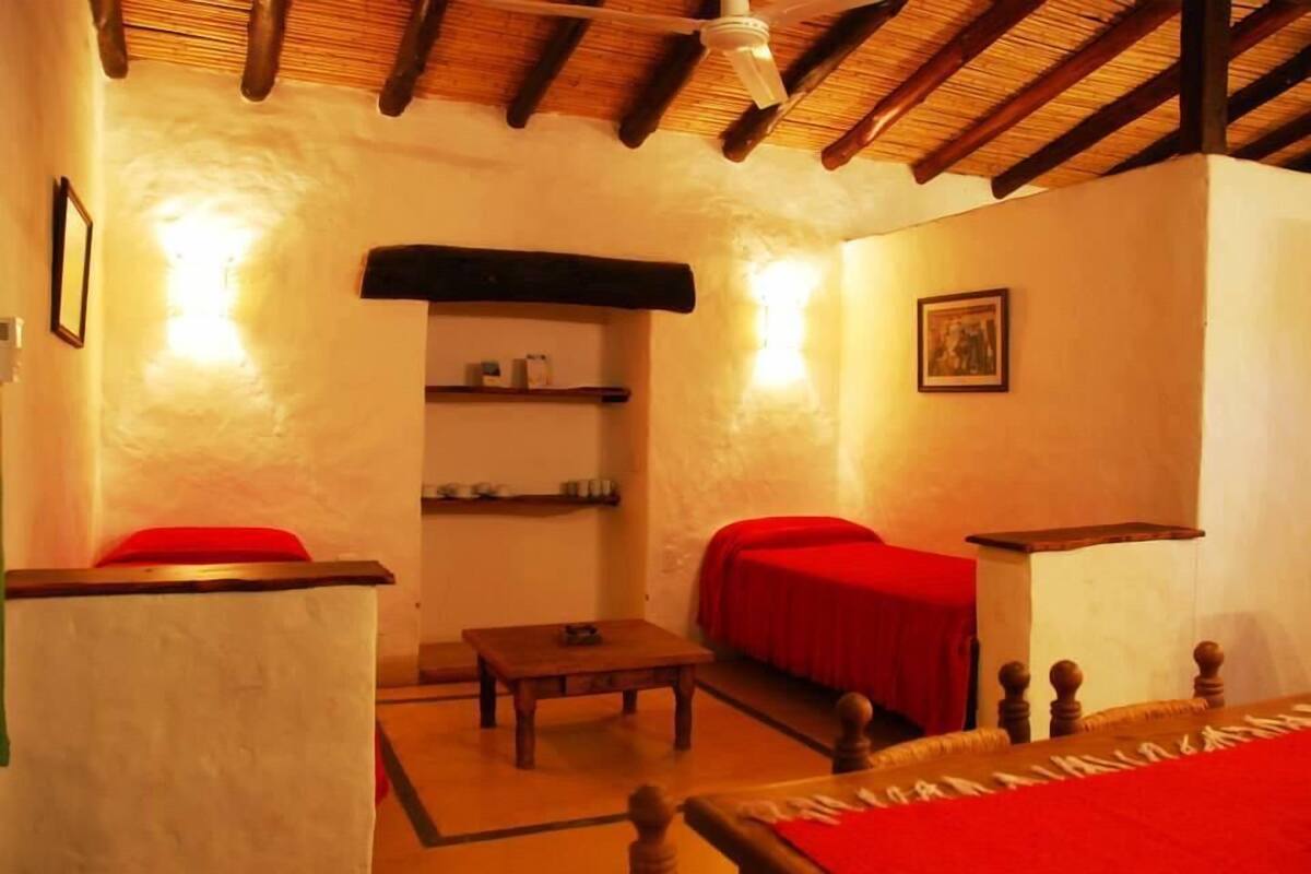 A room with a wooden ceiling, a bed with a red bedspread, a small table, and a shelf with items.
