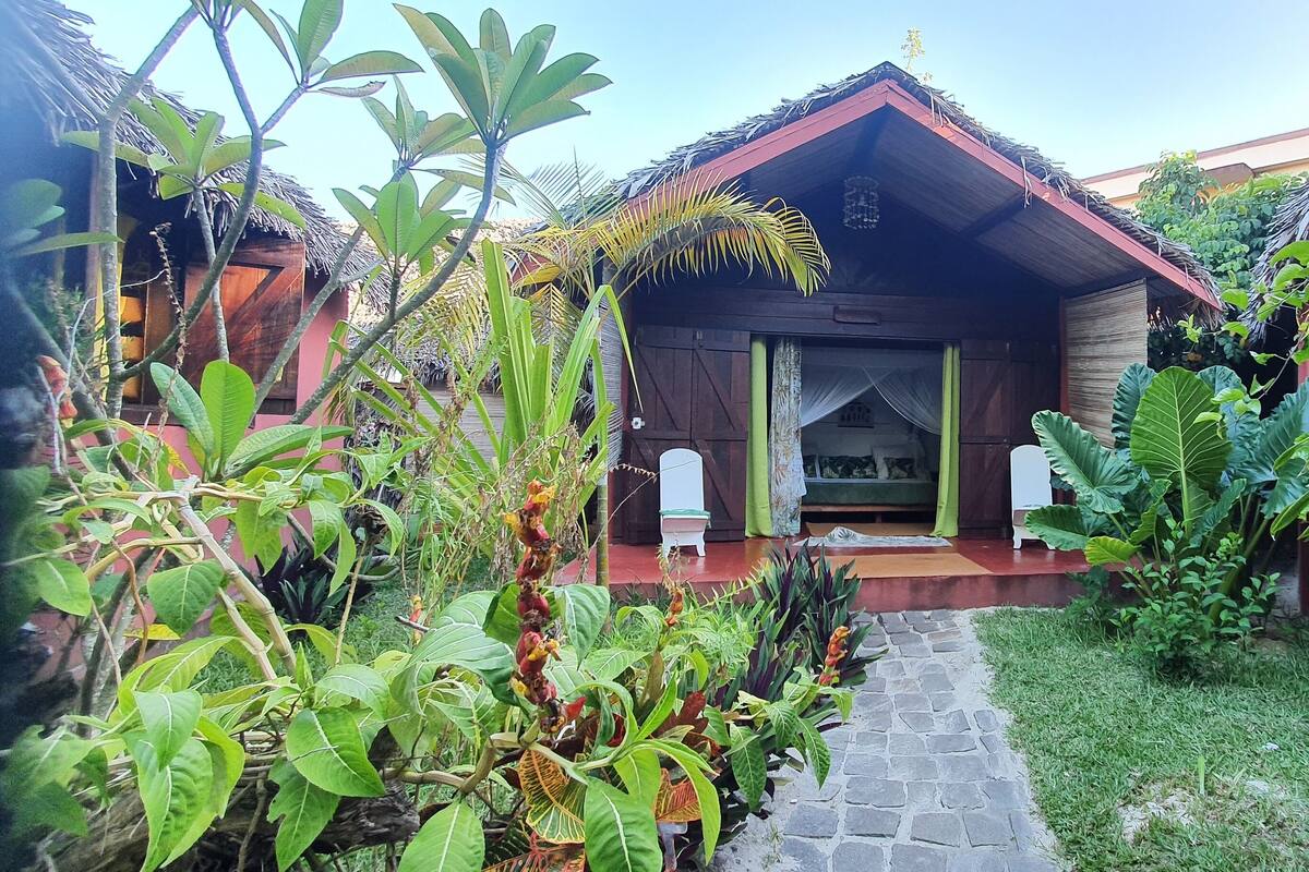 A wooden house with a thatched roof, surrounded by lush greenery and a stone pathway leading to the entrance.
