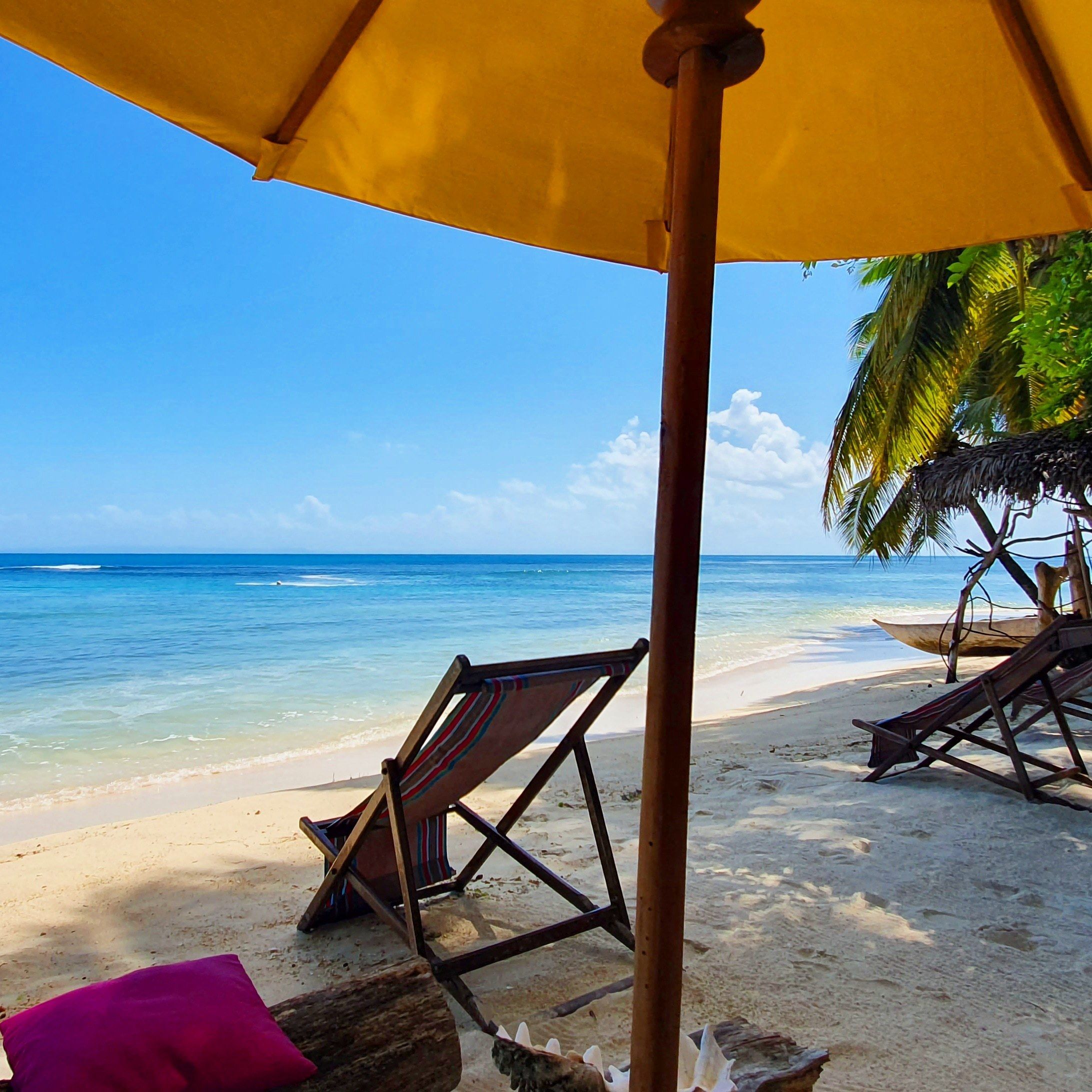 A beach scene with a wooden chair under a yellow umbrella, a wooden bench, and a thatched structure.