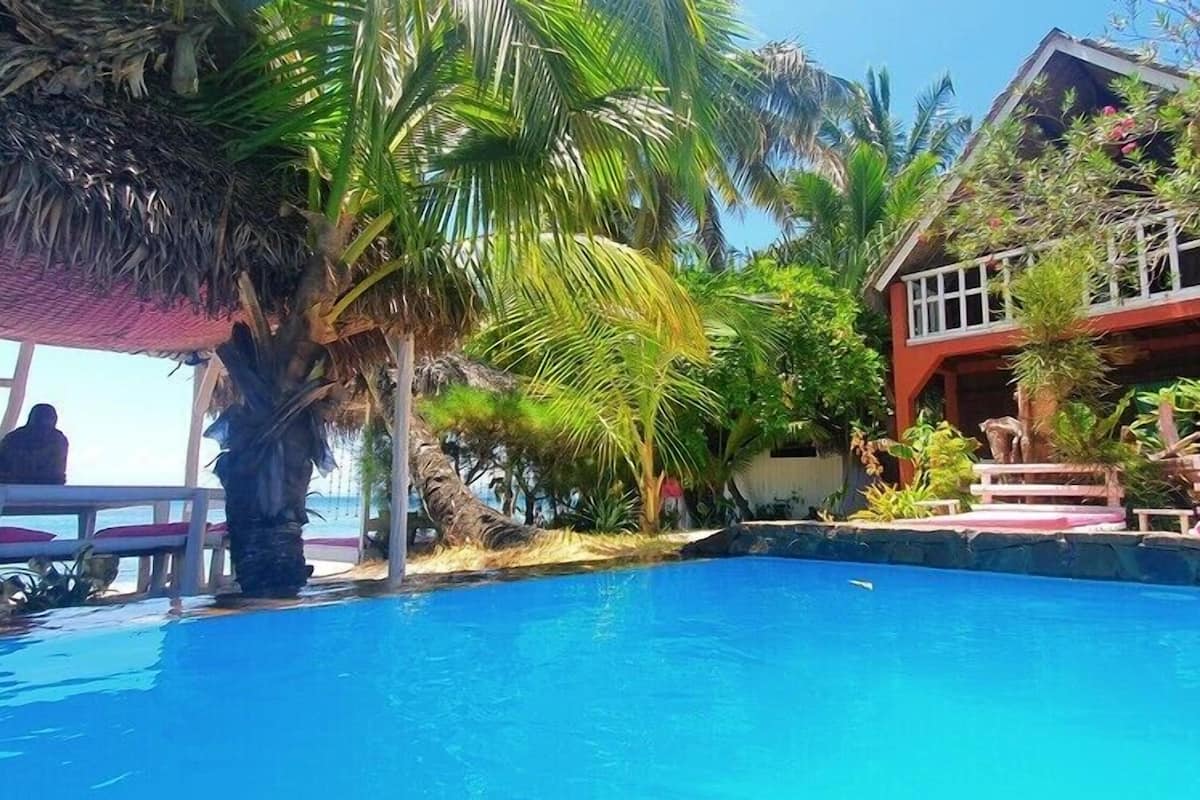 A swimming pool with a thatched roof structure, surrounded by lush greenery and a building with a red facade.