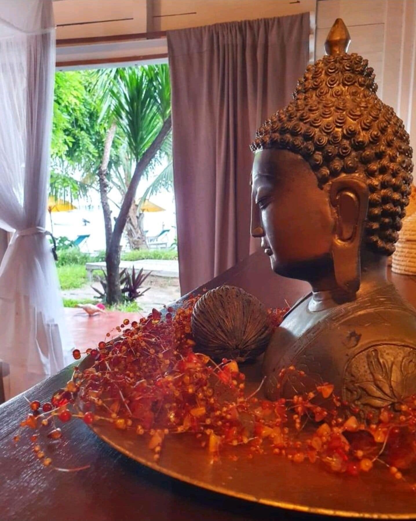 A Buddha statue with a decorative headpiece, surrounded by a tray of red and orange beads, set against a backdrop of a window with curtains and a view of greenery outside.
