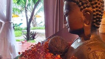 A Buddha statue with a decorative headpiece, surrounded by a tray of red and orange beads, set against a backdrop of a window with curtains and a view of greenery outside.