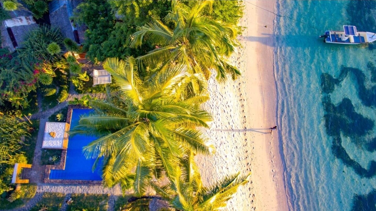 An aerial view of a beach with a boat, palm trees, and a building with a blue roof.
