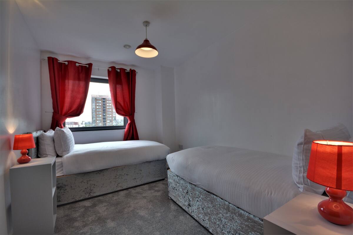 A bedroom with a bed, a window with red curtains, a bedside table with a lamp, and a view of a building outside.