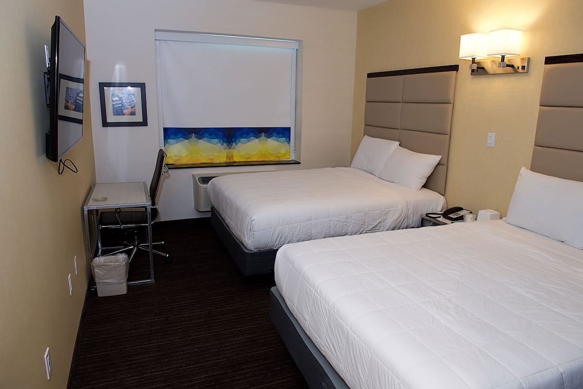 A hotel room with two beds, a desk with a chair, a trash bin, a window with blinds, and a wall-mounted TV.