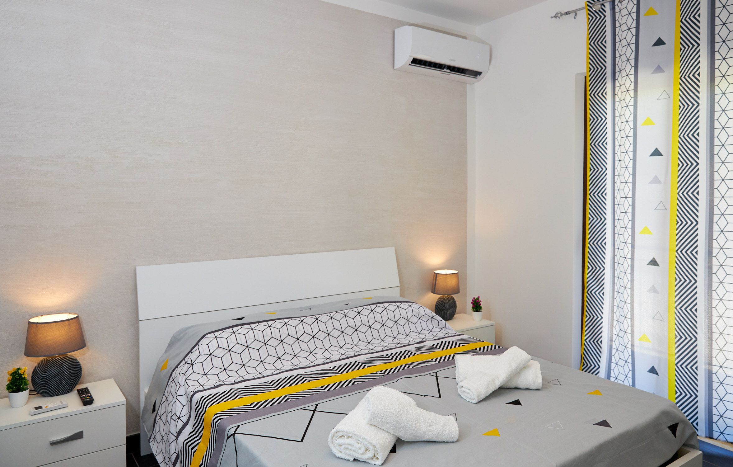 A modern hotel room with a neatly made bed, bedside lamps, a small plant, and an air conditioning unit.