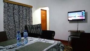 A room with a zebra-patterned curtain, a television mounted on the wall, a table with water bottles, and a patterned tablecloth. - Vosteen Home Holidays (Watamu)