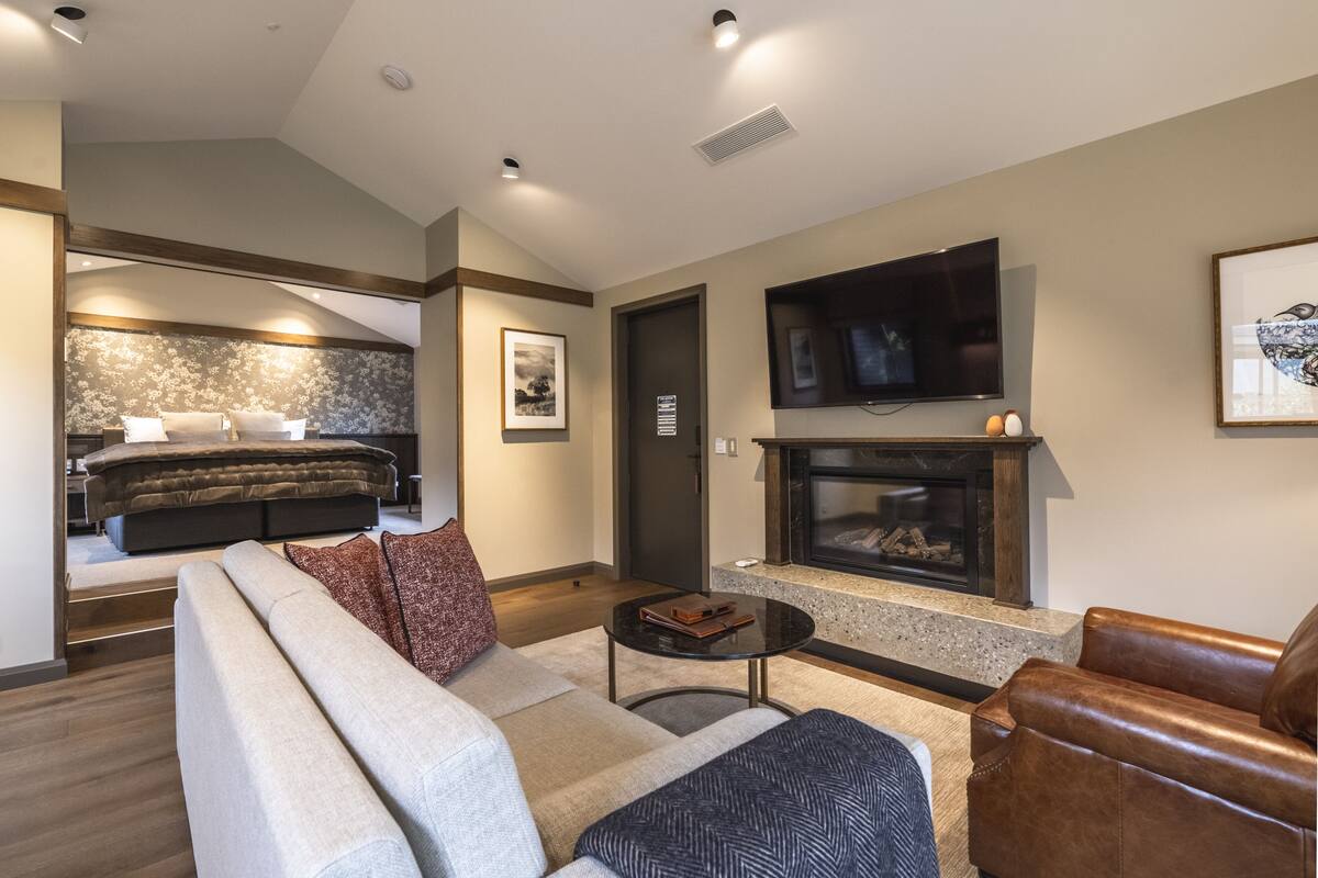 A modern living room with a sofa, coffee table, fireplace, TV, and a bedroom with a bed and nightstand.