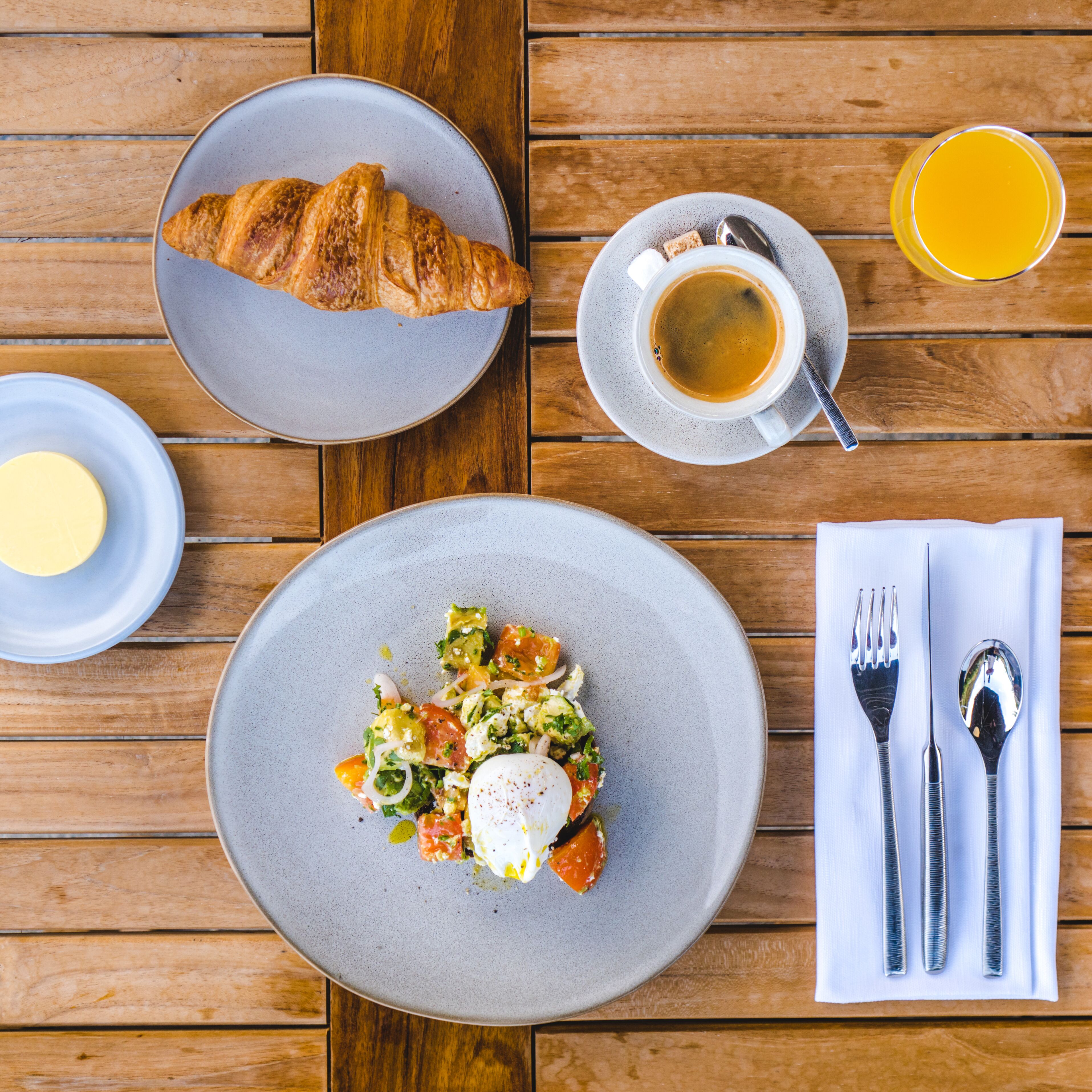 Una mesa con un croissant, una taza de café, un vaso de zumo de naranja, un plato de comida y cubiertos sobre una mesa de madera.