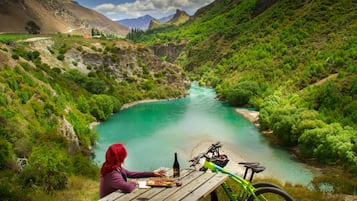 Eine Frau an einem Holztisch im Freien mit Blick auf einen türkisblauen Fluss in einer Berglandschaft.
