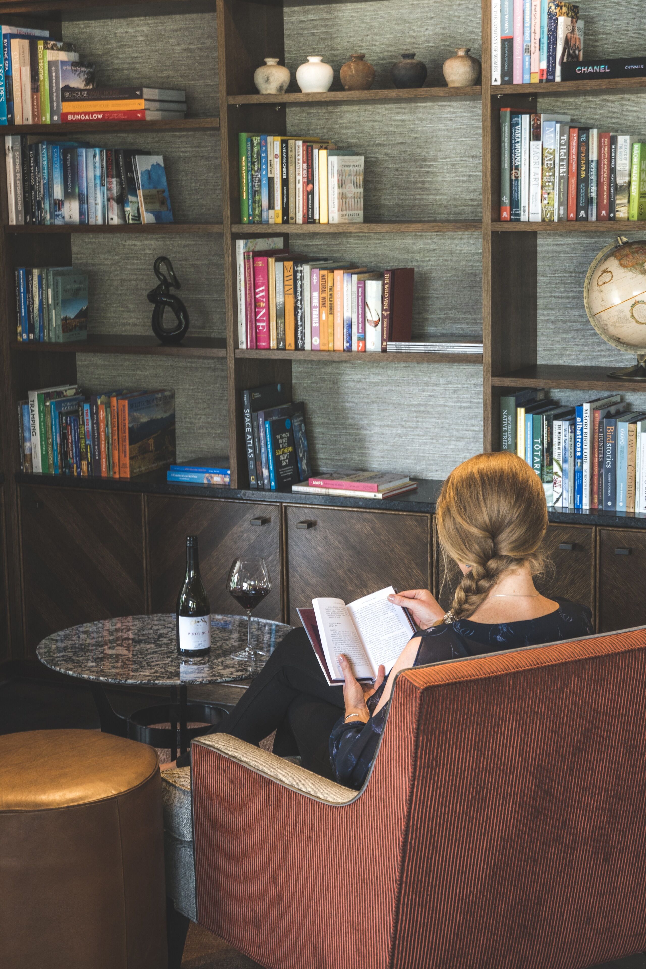 A person reading a book in a room with a bookshelf filled with books, a small table with a bottle and glass, and a round chair.