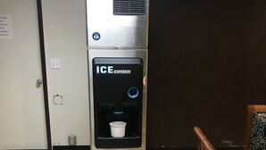 A stainless steel ice dispenser in a room with tiled flooring, a wooden chair, and a door. - Gila Bend Lodge (Gila Bend)