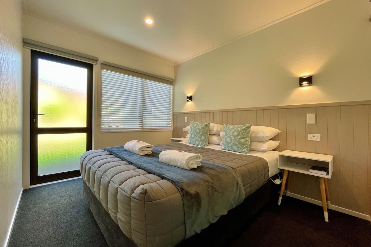 A bedroom with a large bed, a bedside table, and a view of a green landscape through the window.