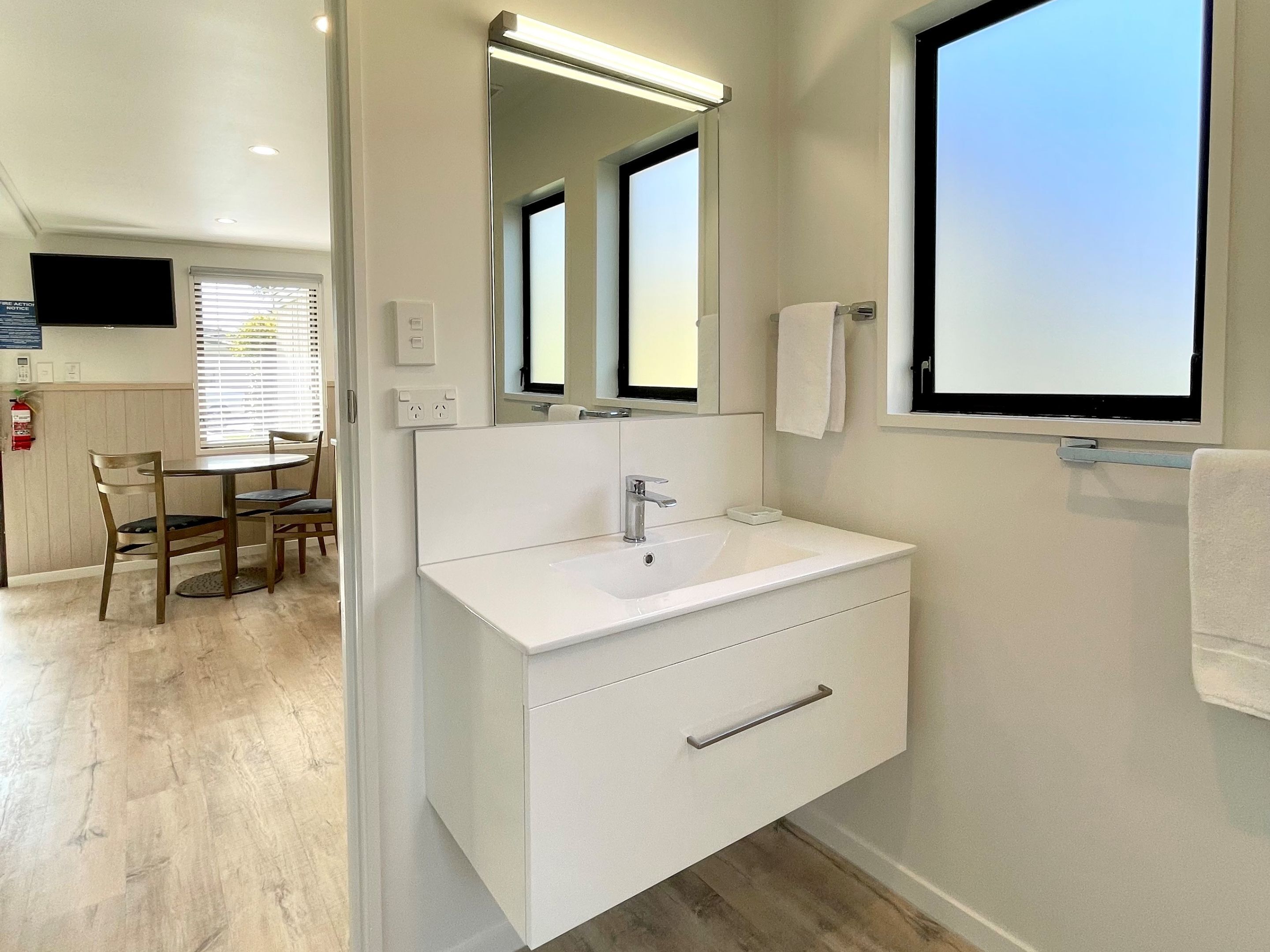 A modern bathroom with a white vanity, a large mirror, and a window. There is a dining area visible in the background.