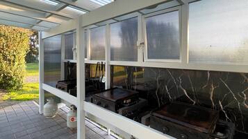 Cocina al aire libre techada con estufas a gas y una parrilla, bajo una pérgola blanca con ventanas de malla metálica.