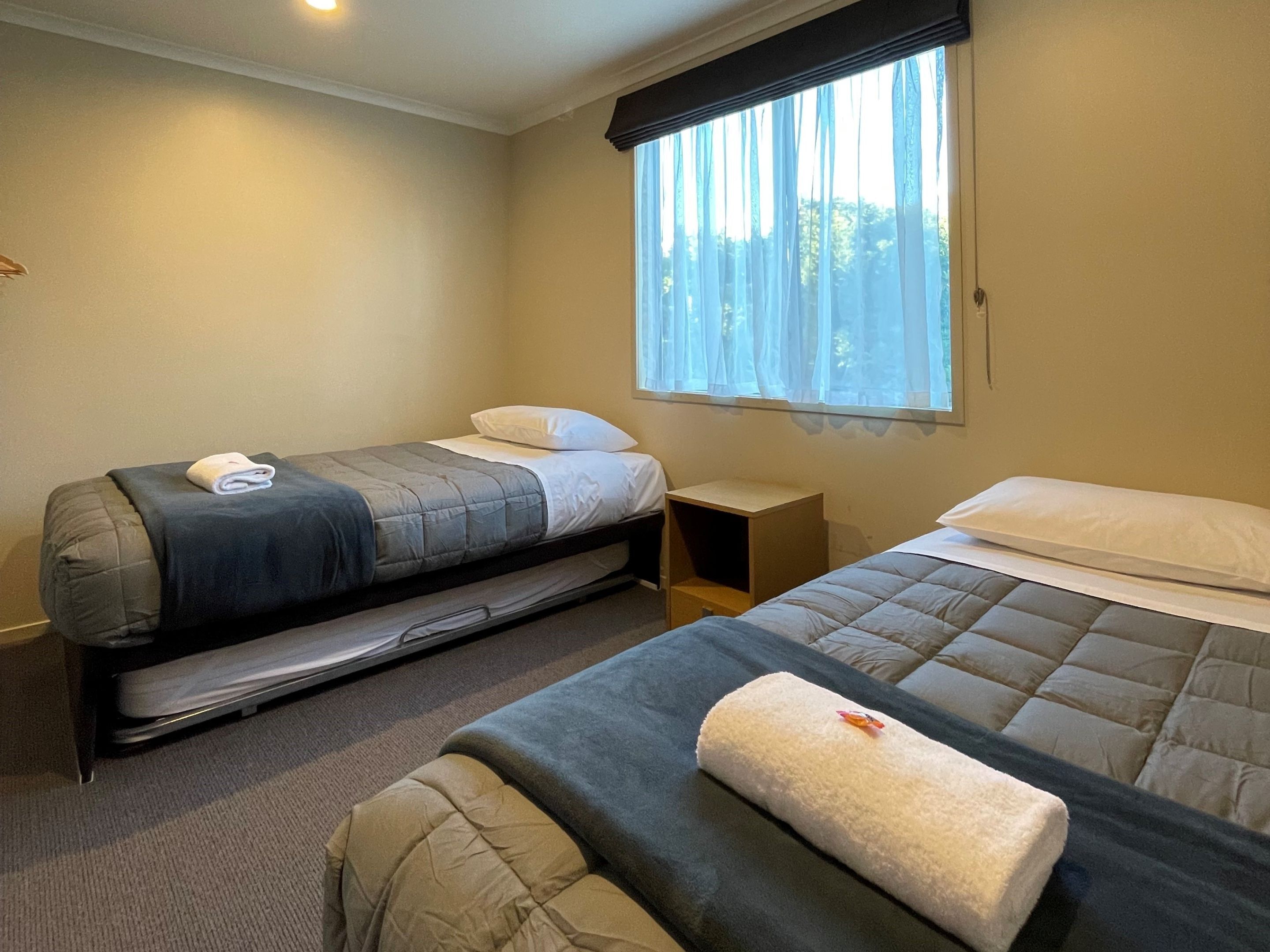 Una habitación con dos camas, una mesita de noche de madera y una ventana con cortinas ligeras.
