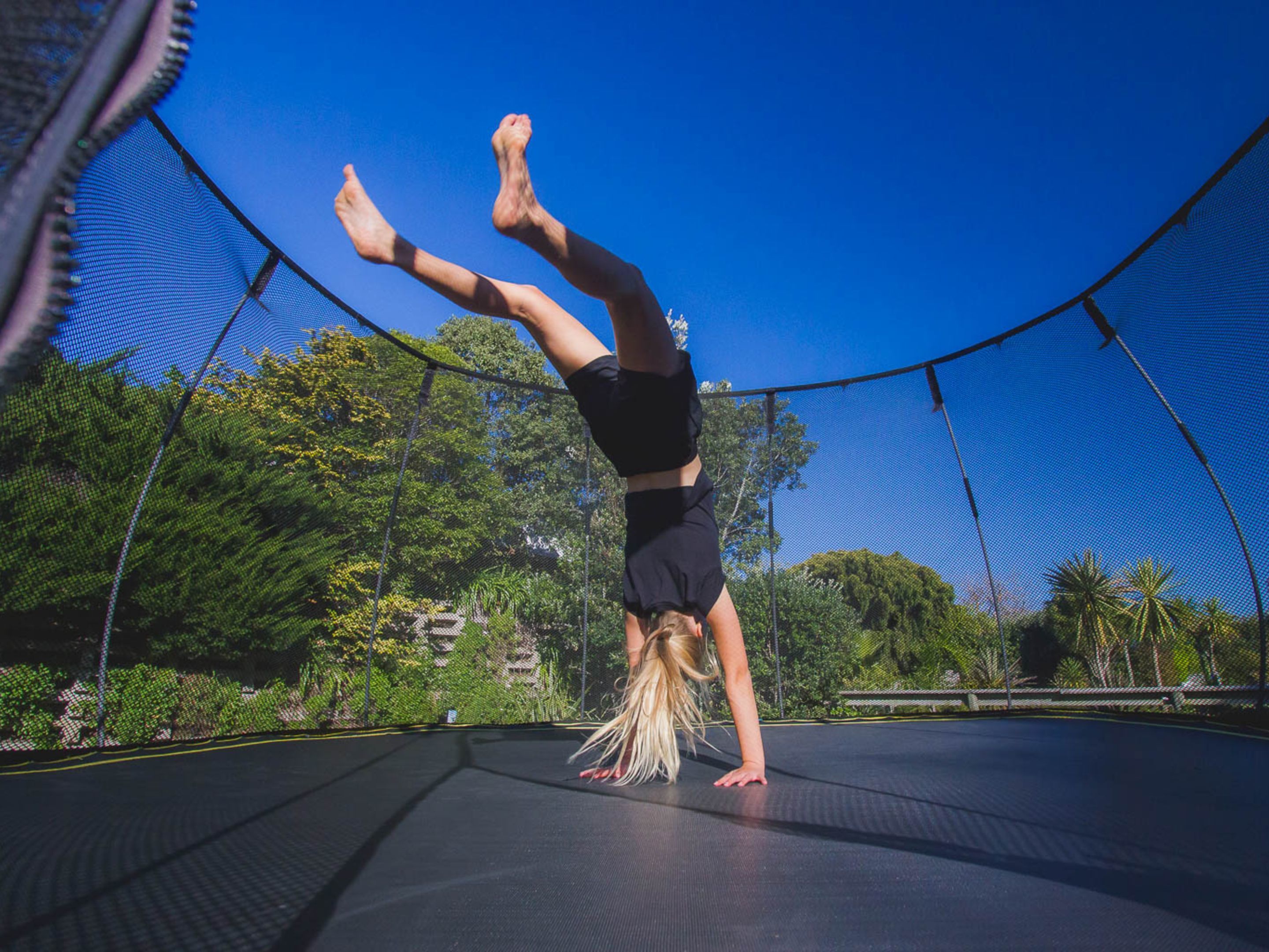 Una persona che fa la verticale su un trampolino circondato da una rete di sicurezza, con alberi e un cielo sereno sullo sfondo.