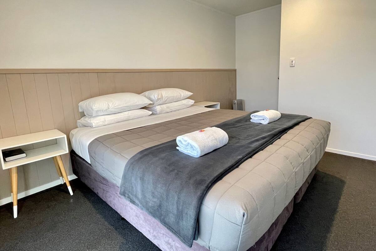 A neatly made bed with pillows and a blanket, a bedside table with a lamp, and a power outlet on the wall.