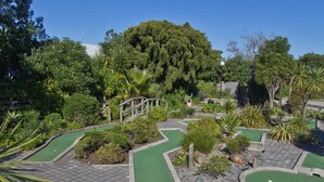 A miniature golf course with multiple green putting areas, a wooden bridge, and surrounding lush greenery. - Pacific Park Christian Holiday Camp (Tauranga)