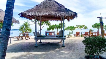 Un espacio de relax frente a la playa, con techo de paja, sillas de madera y una pequeña mesa.